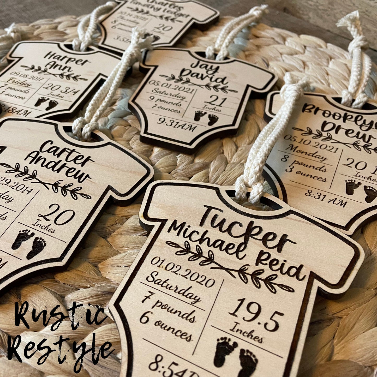 6 natural wooden ornaments shaped like baby onesies and hung by white cotton braided strings laying flat on a table. These items are engraved with each baby's name and birth stats.