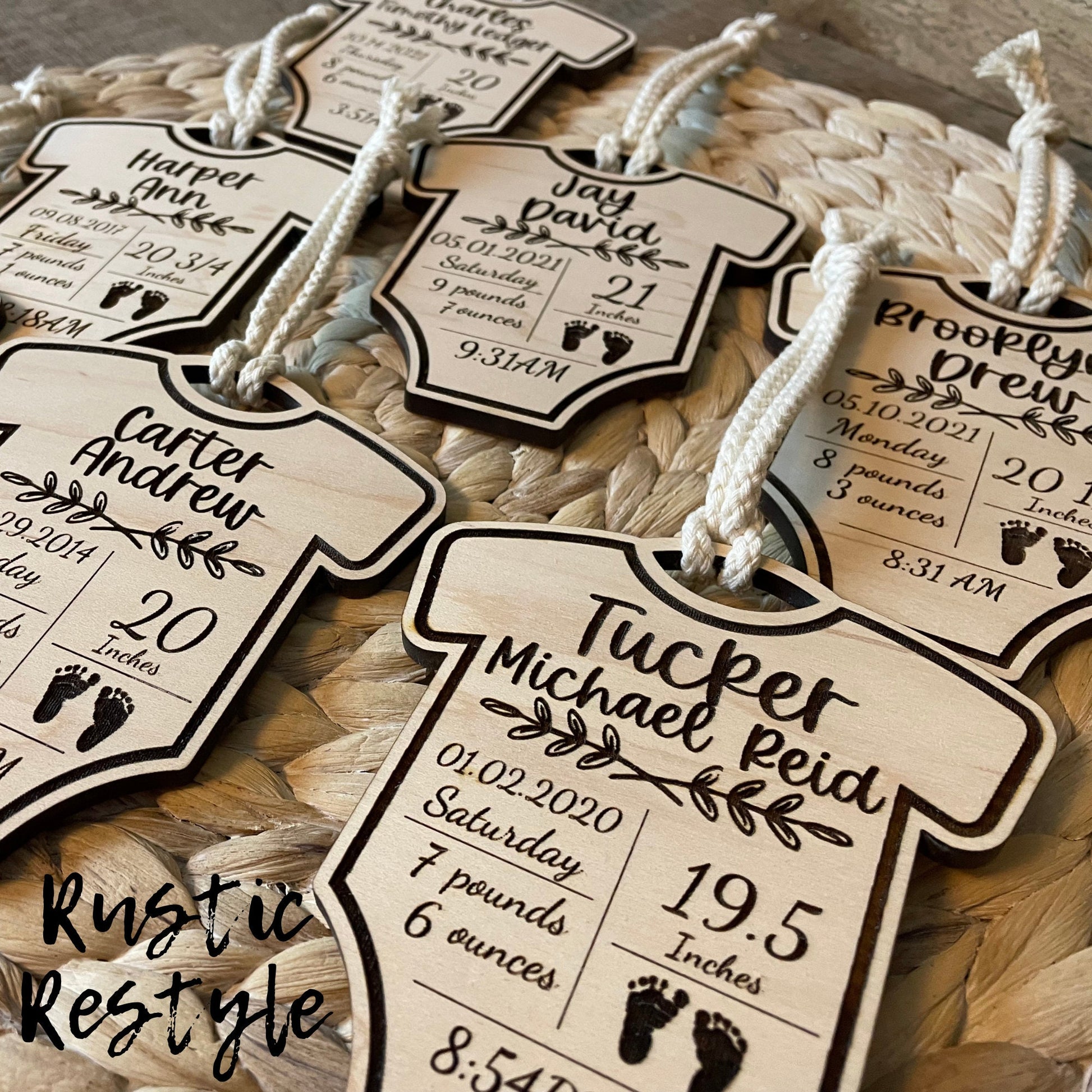 6 natural wooden ornaments shaped like baby onesies and hung by white cotton braided strings laying flat on a table. These items are engraved with each baby's name and birth stats.