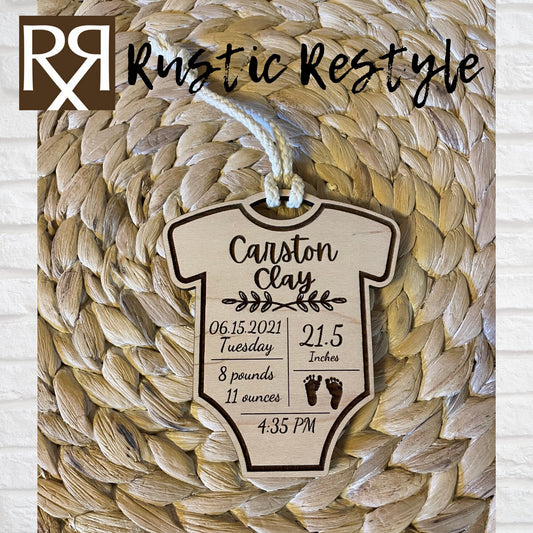 A natural wooden ornament shaped like a baby onesie and hung by a white cotton braided string. This Item is engraved with the baby's name and birth stats.