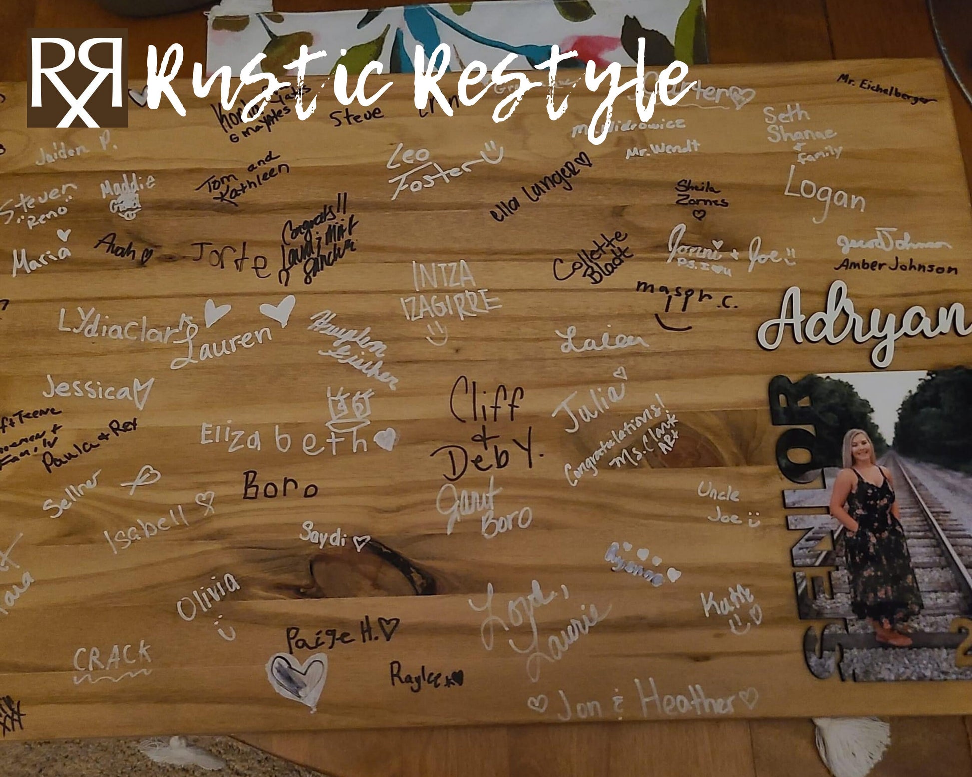 Graduation Guestbook, Large wood rectangle board laying flat on a table. The board has a senior photo in the lower right corner with the name Adryan above it.The board is covered in black and white signatures.