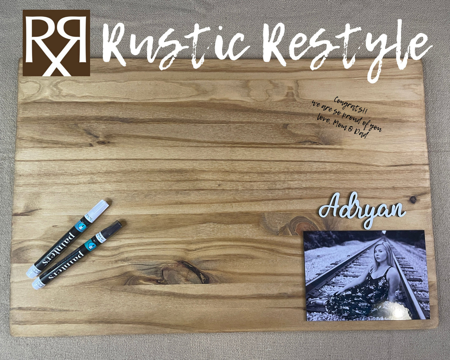 Graduation Guestbook, Large wood rectangle board laying flat on a table. The board has a senior photo in the lower right corner with the name Adryan above it. There are Markers for signing lying on the board with an example signature in black.
