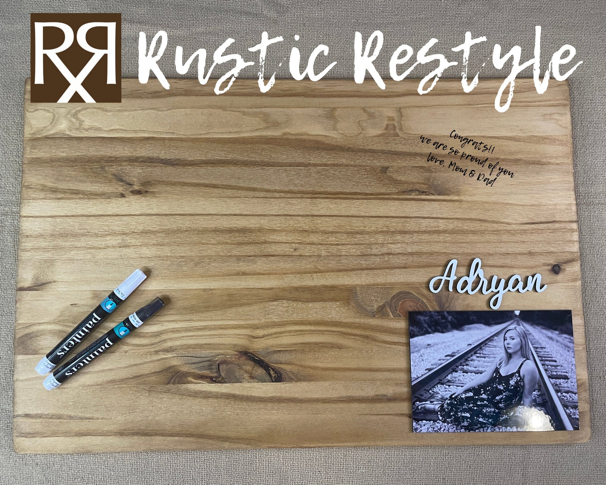 Graduation Guestbook, Large wood rectangle board laying flat on a table. The board has a senior photo in the lower right corner with the name Adryan above it. There are Markers for signing lying on the board with an example signature in black.