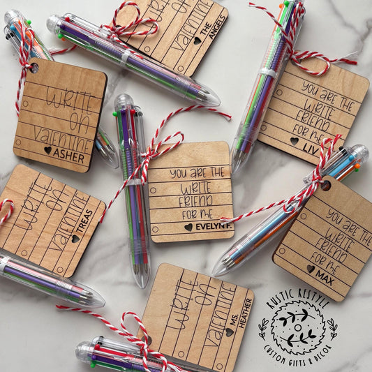 “Personalized wooden Valentine’s Day tags paired with multicolor click pens, featuring engraved messages like ‘You are the write kind of friend’ and ‘Write on Valentine,’ each tied with string and customized with a child’s name.”