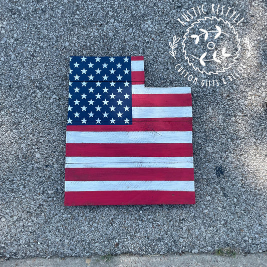 Wooden Utah‑shaped pallet sign painted as an American flag with stars and stripes, rustic finish, displayed on outdoor concrete.