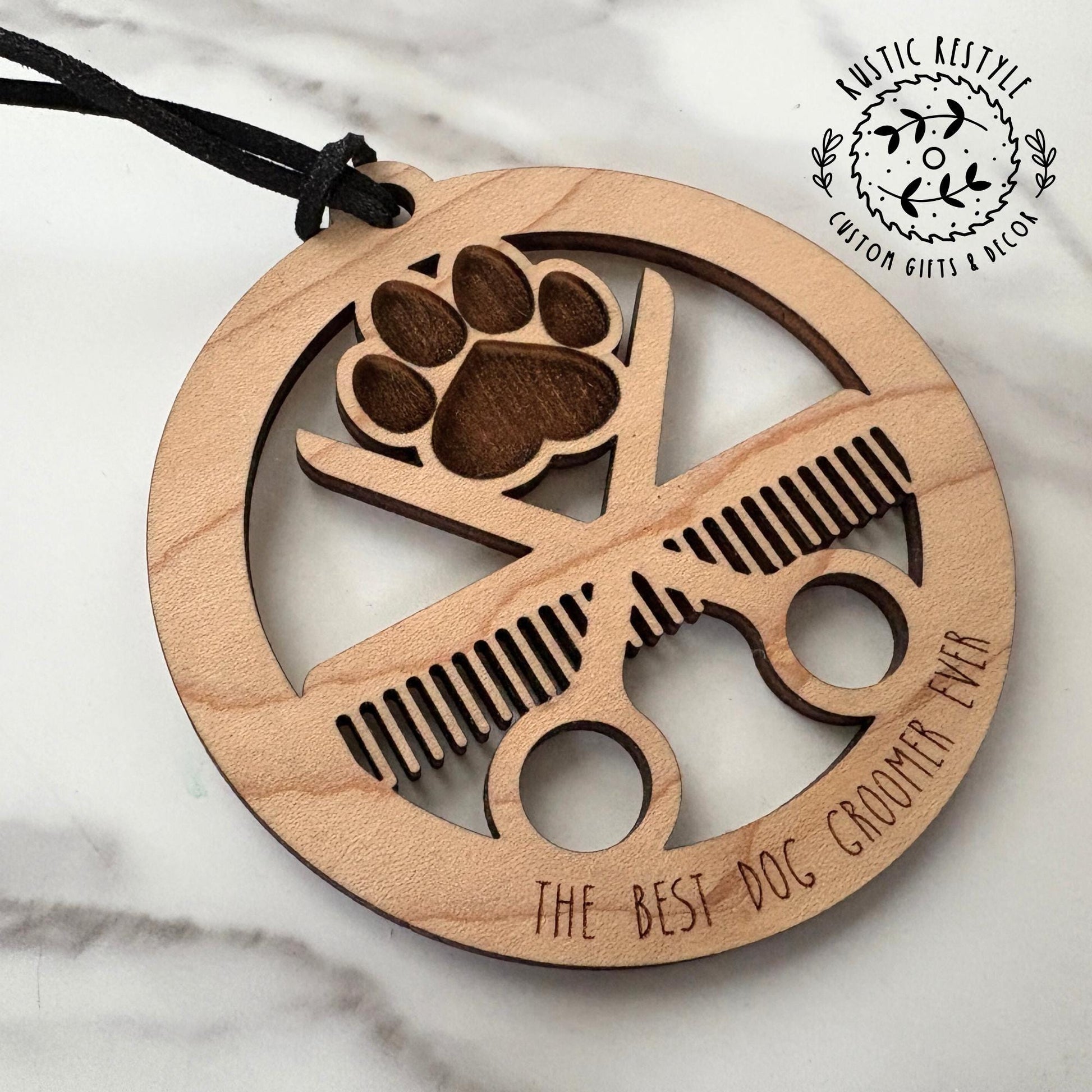Wooden dog groomer ornament featuring a circular design with laser-cut grooming scissors, comb, and paw print. Engraved with the phrase "THE BEST DOG GROOMER EVER" and hung by a black cord. Rustic style suitable for holiday or year-round display.