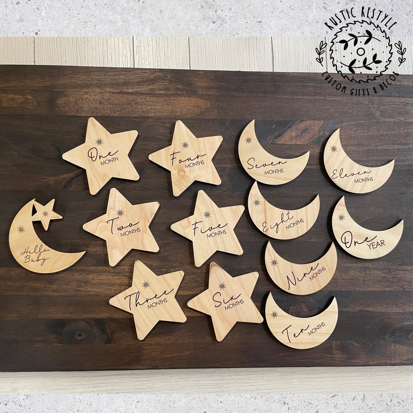 Wooden star and moon baby milestone markers arranged on a dark wood surface, engraved with monthly milestones from Hello Baby to One Year.