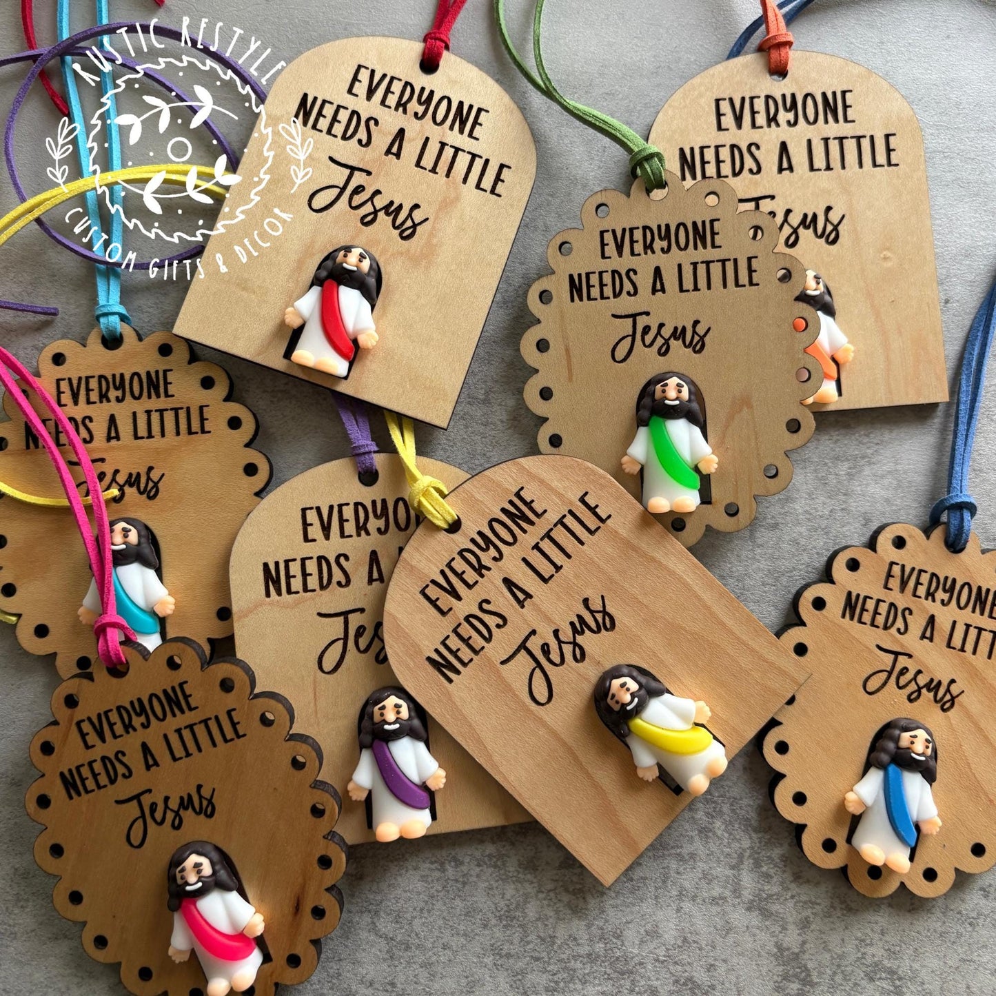 Handmade wooden gift tags featuring small Jesus figurines and engraved text “Everyone Needs a Little Jesus,” each with a colorful string loop.