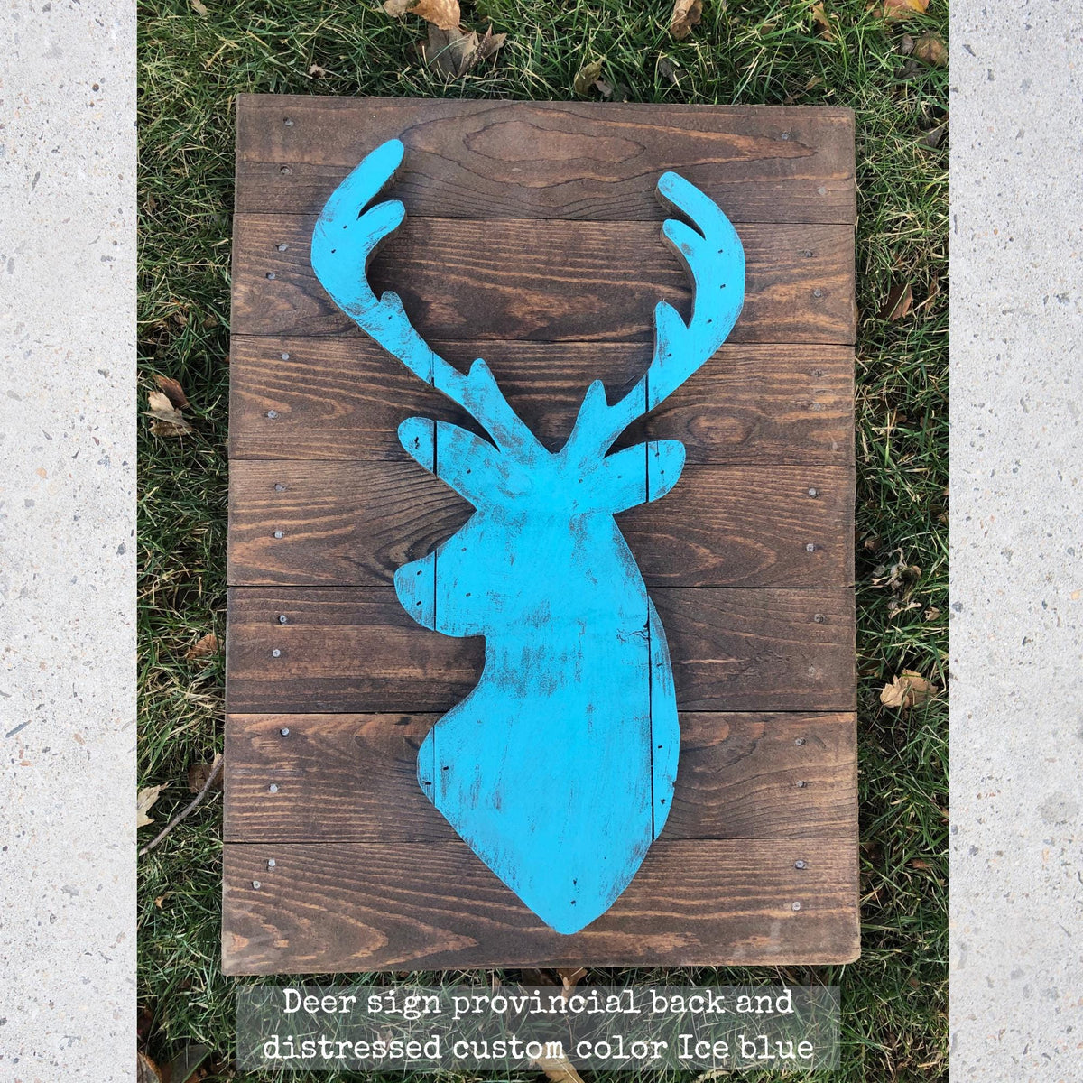 A wooden deer head sign with a distressed blue color, placed on a wooden plank against a grassy background.