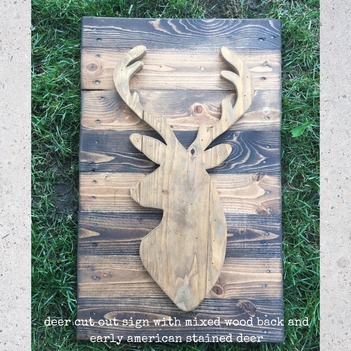 A wooden deer head cut out sign with a mixed wood back and early American stained deer, placed on a wooden board against a grassy background.