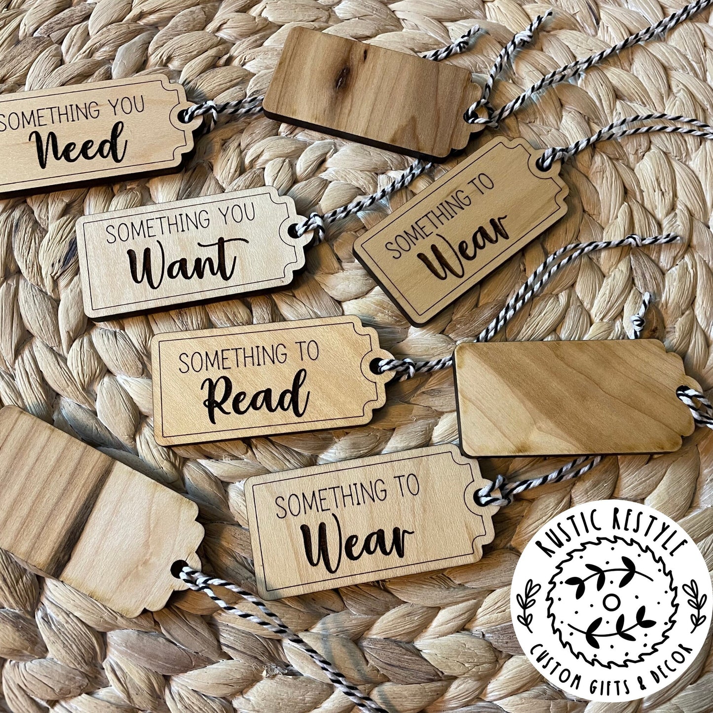 A pile of Christmas gift tags laying on a woven placemat. A mix of 4 sayings, Something to : Want, Wear, Read, and Need. Tags show both fronts and backs of tags. Each tag has a black and white bakers twine string.